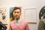 A person in a light pink cardigan and white shirt stands before a gallery wall showcasing Steve Kims vibrant artworks, which blend colorful abstract creations with striking monochrome pieces, highlighting the exhibitions eclectic charm.