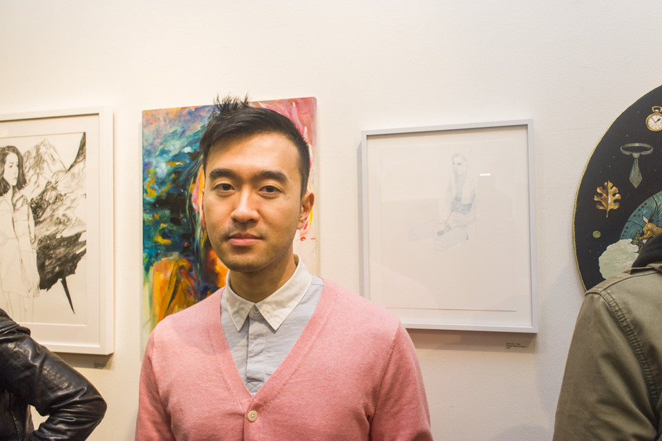 A person in a light pink cardigan and white shirt stands before a gallery wall showcasing Steve Kims vibrant artworks, which blend colorful abstract creations with striking monochrome pieces, highlighting the exhibitions eclectic charm.