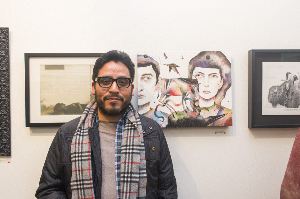 A person wearing glasses and a plaid scarf stands in front of a wall with framed artwork, featuring a colorful gouache abstract painting and black-and-white illustrations by Ken Garduno on printmaking paper.