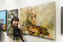 A woman stands next to a wall displaying various artworks, including Soey Milks oil on canvas. The central piece features a woman gazing at an antler in her hand, wrapped in a shawl and seated on a patterned blanket. Art for sale is available throughout the gallery.