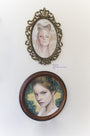 Two framed artworks grace the white wall: above, an oval brass frame features a woman with a headband in Bec Winnels style; below, Genevieve is captured in a round wooden frame with an intricate hairstyle set against a green-yellow background.