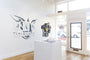 In a bright art gallery featuring white walls and wooden floors, Megan Wolfes Wings & Pieces #15 is displayed among abstract designs and paintings on the left wall. A small pedestal with pamphlets stands centrally, while large windows and a glass door enhance the right side.