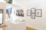 A small art gallery interior is adorned with framed portrait paintings and sketches on white walls. Through the open entry door, a person admires the display, likely attracted by a piece from Michael Ramstead. In the center, a table holds booklets and flyers for upcoming exhibitions.