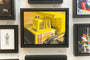 Brand Name artist Allan Innman presents a framed oil on canvas showcasing a close-up view of a yellow Tonka toy dump trucks side, with the iconic logo vividly featured among other artworks.