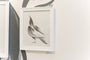 The framed artwork Wings & Pieces #15 by Megan Wolfe displays a monochrome abstract bird with flowing lines on a light background. Its rendered in pencil on paper, set in a white frame, and encased in clear glass.