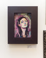 A framed painting by Rich Pellegrino, titled Hope, features a woman with long hair and closed eyes in abstract purple, blue, and pink. The dark wood frame adds depth. A label below reads: Hope, 5x7, acrylic on board, $500, currently showcased at Spoke Art Gallery.