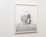 A framed artwork by Jonas Lofgren on a white wall depicts a graphite on paper drawing of a house with dual roofs, ornate facade, and an antenna-like structure, set against an abstract textured landscape.