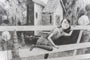 This Jonas Lofgren piece in graphite on paper shows a short-haired girl in a dress, leaning on a wooden fence with one leg raised. A small house, trees, and abstract shapes form the backdrop. The elegantly framed artwork enhances the mysterious scene in black and white.