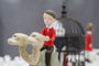 A hand-sculpted resin figurine from Doubleparlour, featuring a person with short brown hair in a red shirt, perches on a whimsical two-headed camel. An out-of-focus metal birdcage provides an intriguing backdrop to this acrylic-painted scene.