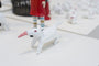 A small white ceramic figure by Doubleparlour, featuring a red cone-like nose and possibly hand-sculpted resin, stands on a flat surface. Similar figures and the lower part of a larger striped-stocking figure in the background create an eclectic scene.