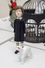 A resin sculpture by Doubleparlour features a young girl in a black dress with white collar, green-striped socks beside a small white creature. An open black wire birdcage adds intrigue, all set on a smooth white surface, varnished and painted with acrylics.
