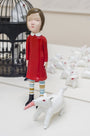 A Doubleparlour hand-sculpted girl in a red dress, striped leggings, and black shoes stands by a whimsical white animal with a carrot-shaped horn. Vivid acrylics adorn the figures. In the background are a birdcage and more small white animals.
