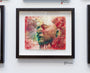 A framed artwork by Akira Beard displays a painted human face with closed eyes, merging vibrant red, green, and blue inks. Text adorns the face, and it is set against a white wall alongside other framed pieces.