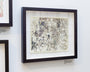 A framed Barron Storey-inspired piece hangs on a white wall, showcasing an intricate abstract line drawing. The dark wood frame includes a small, unclear label below the image. Ink and acrylic add depth to the artwork.