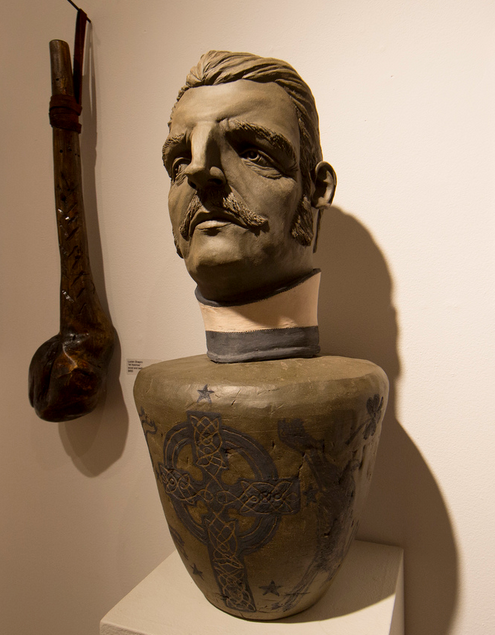 A ceramic artwork by Derek Weisberg portrays a man with a mustache and traditional attire atop an urn with Celtic designs. In the background, a carved wooden club adds homage, subtly reminiscent of a Martin Scorsese tribute.