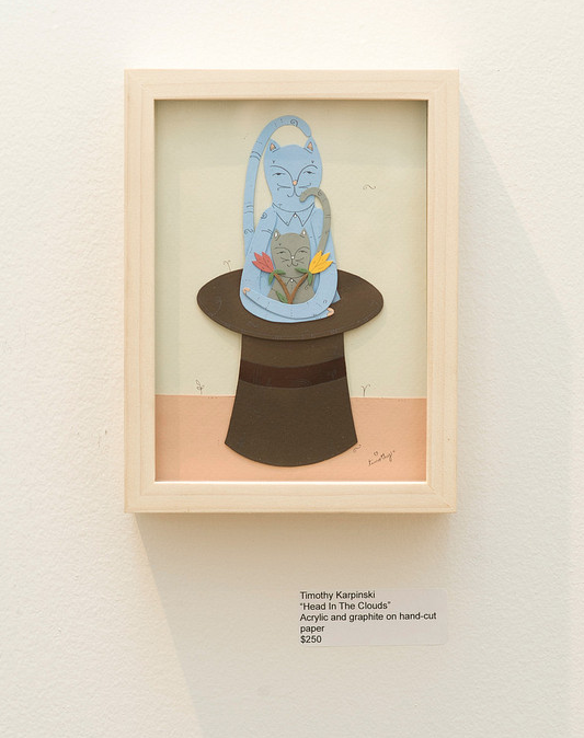 A hand-cut paper artwork by Timothy Karpinski features a whimsical, blue-hued cat on a top hat surrounded by vibrant flowers. The plaque says In The Clouds, created with acrylic and graphite, priced at $250.