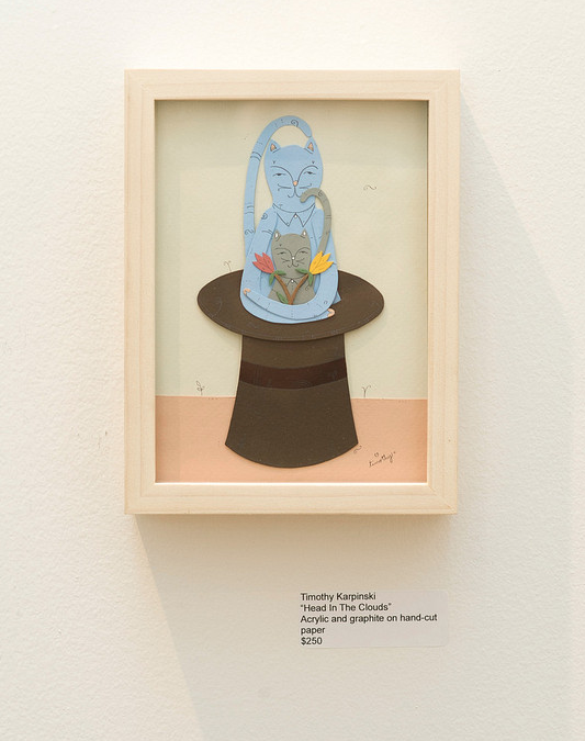 A hand-cut paper artwork by Timothy Karpinski features a whimsical, blue-hued cat on a top hat surrounded by vibrant flowers. The plaque says In The Clouds, created with acrylic and graphite, priced at $250.
