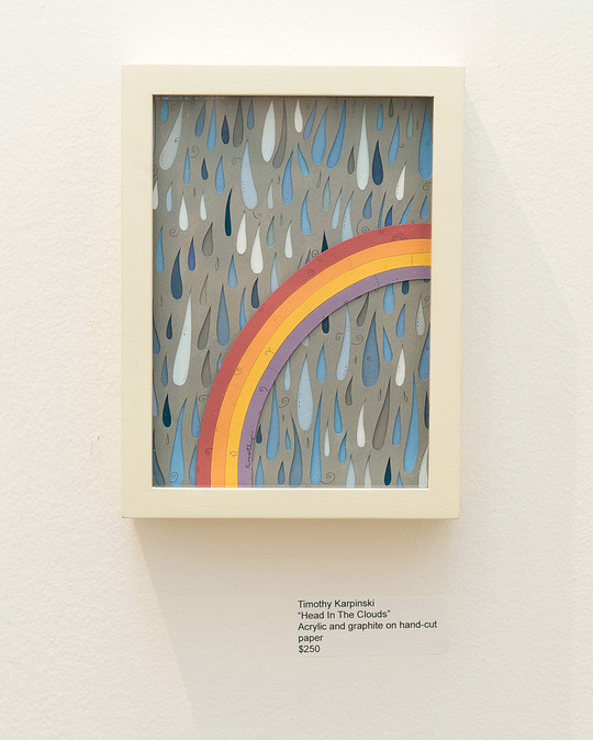 A framed artwork by Timothy Karpinski, titled Head in The Clouds, features a rainbow with stylized raindrops on a cloudy background and is on display at Spoke Art Gallery. It uses acrylic and graphite on hand-cut paper and is priced at $250.