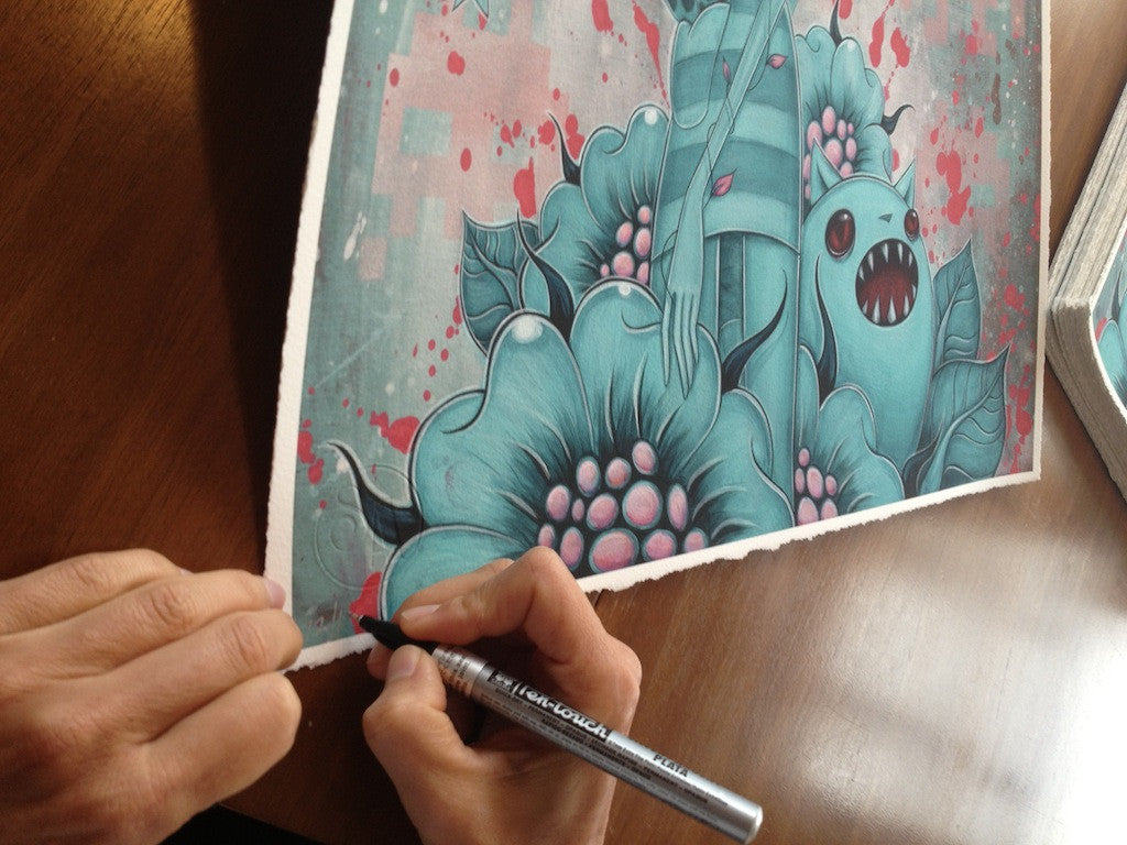 A person uses a silver marker to sign and number a limited edition Caia Koopman fine art giclee print, showcasing cartoonish blue flowers and a small teal creature with an open mouth. It rests on the wooden table, poised for its spot in an exclusive collection.