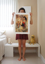 In a bedroom, a person holds a limited edition Soey Milk painting showing a woman with abstract elements in front of their face. Theyre barefoot, wearing a red skirt on the white floor near a bed and bench adorned with a yellow crochet figure.