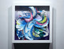 Crystal Wagners vibrant abstract art showcases layered swirling shapes in blue, pink, green, and white. The textured design resembles waves or feathers and is framed in a white square frame on a wall, reflecting cut paper techniques.