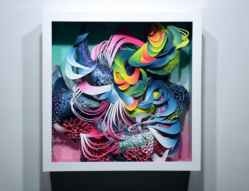 A white-framed abstract paper sculpture by Crystal Wagner features swirling layers of vibrant pink, blue, yellow, and green colors with intricate textures creating a 3D relief effect.