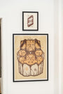 Two framed artworks adorn the white wall. The larger GATS piece, a signed and numbered edition, features an abstract face with geometric patterns in brown and beige. Above it, a smaller frame displays two stylized feathers on a brown background, echoing an Infinite Eye design.