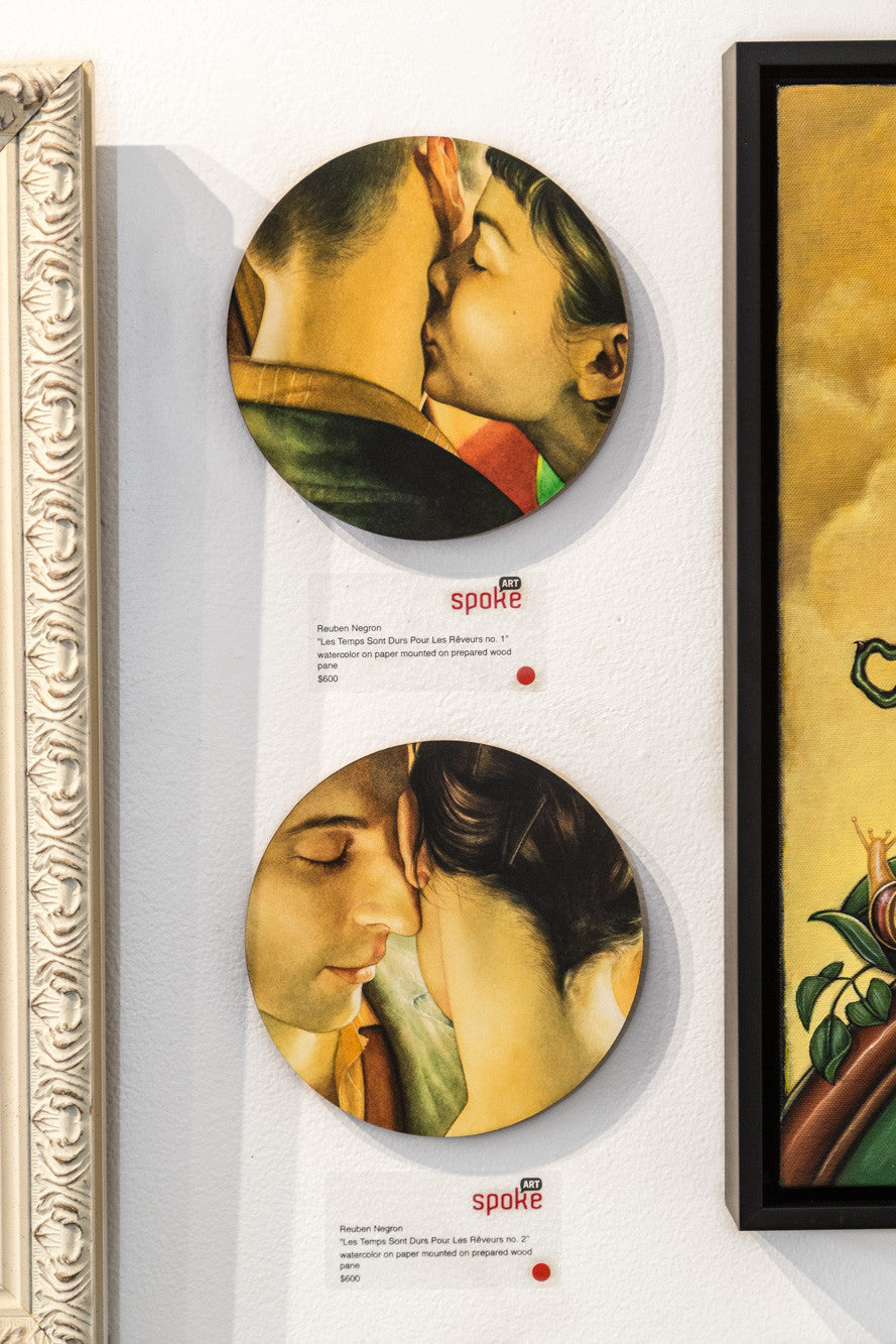 Two circular watercolor artworks by Reuben Negron depict intimate moments: the top shows a gentle kiss on the forehead, while the bottom features tenderly touching heads. Small labels beneath each piece detail their sale, enhancing their allure.