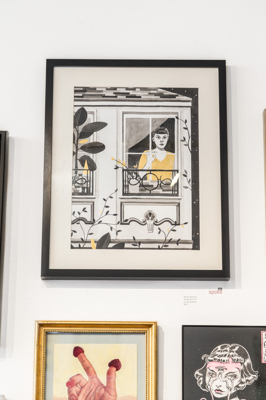 Monica Garwoods framed artwork features a person with dark hair and a yellow shirt, gazing through an ornate window with vibrant plants. This black and white illustration is reminiscent of Amelie Sur le Fil and is displayed with other striking pieces.