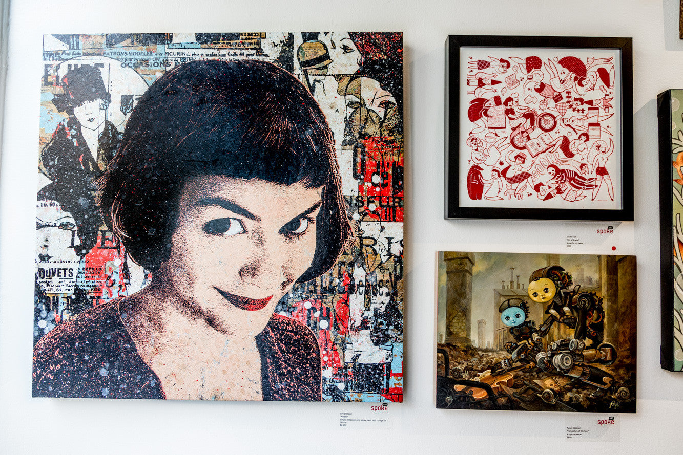 A wall displays three artworks: a textured portrait of a smiling woman with short hair like Amélie, an abstract red and white illustration, and Greg Gossels acrylic steampunk-style painting featuring robot characters.
