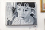 A Rebecca Adams acrylic on canvas, featuring a thoughtful woman with short hair and overlaying white abstract lines, is elegantly showcased in a light-colored frame on a gallery wall.