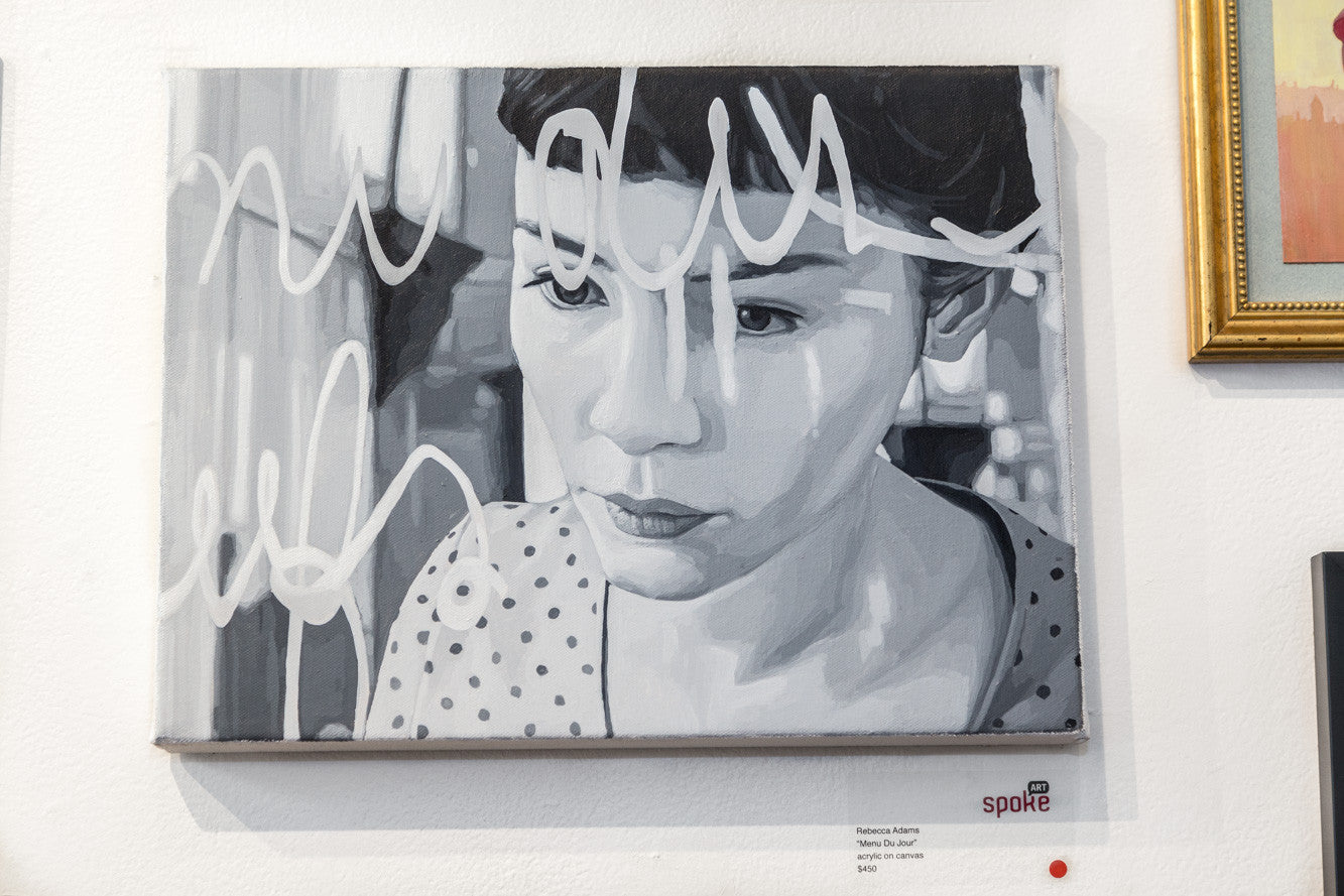 A Rebecca Adams acrylic on canvas, featuring a thoughtful woman with short hair and overlaying white abstract lines, is elegantly showcased in a light-colored frame on a gallery wall.
