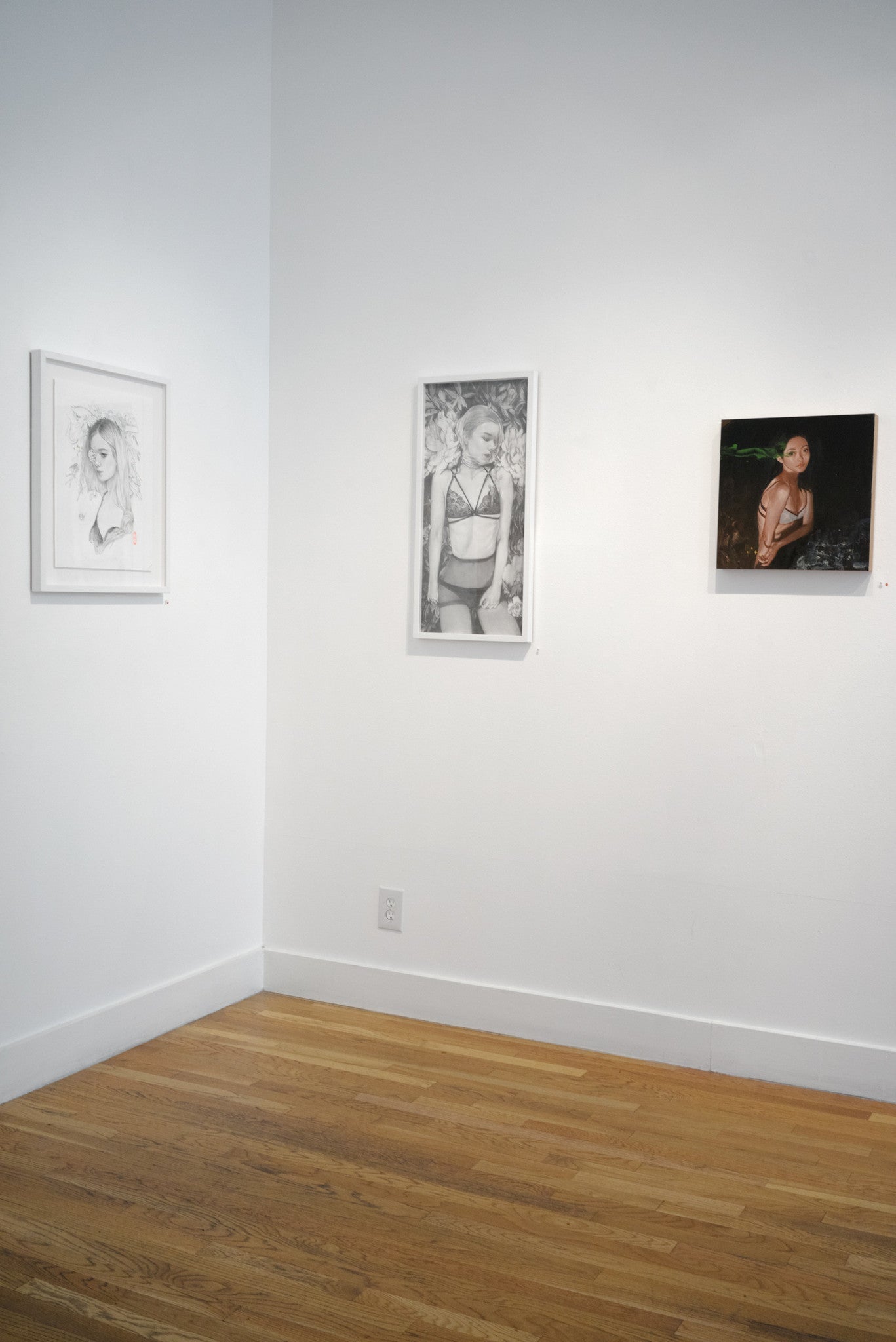 A white gallery wall displays three artworks: a sketch of a woman, a black-and-white floral-themed photograph of a woman, and Helice Wens acrylic on wood painting showing a woman in shadow. The setting’s elegance is elevated by the warm wooden flooring.