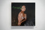 A Helice Wen painting showcases a woman in a bra with crossed arms against a dark backdrop, highlighted by a green brushstroke near her face evoking Northern Light. This acrylic on wood panel features abstract, smoky textures and is displayed on a white wall.
