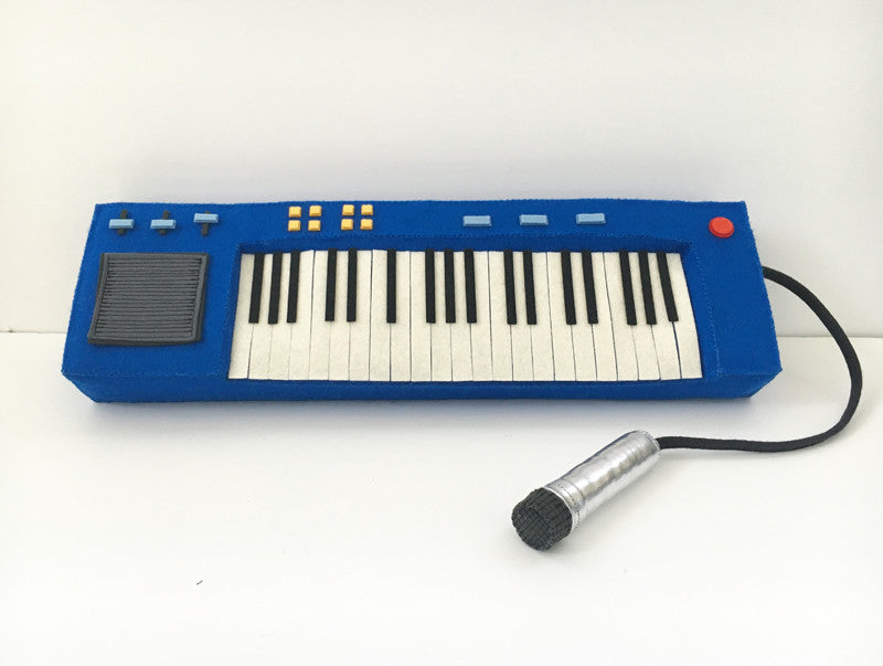 Steff Bombs blue felt toy keyboard, inspired by Bobs Burgers art, features black and white keys, control buttons, and a display. A black cord connects it to a silver and black felt microphone, all set for Gallery1988 fans on a white surface.