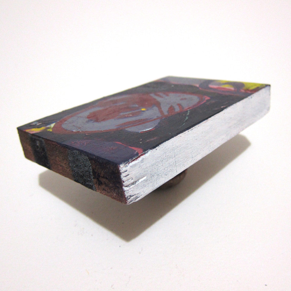 This abstract art piece by Tessar Lo features a painted image on a rectangular block with vibrant red, yellow, and black colors. Elegantly resting on a textured wooden base against a white background, it captures the essence of mixed media artistry.