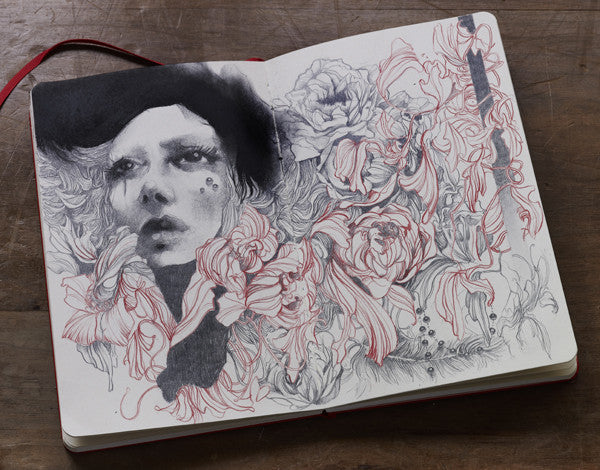 The Craww open sketchbook displays an expressive face surrounded by blooming flowers in grayscale and red tones. This graphite artwork blends seamlessly with the floral elements, set against a wooden surface background.
