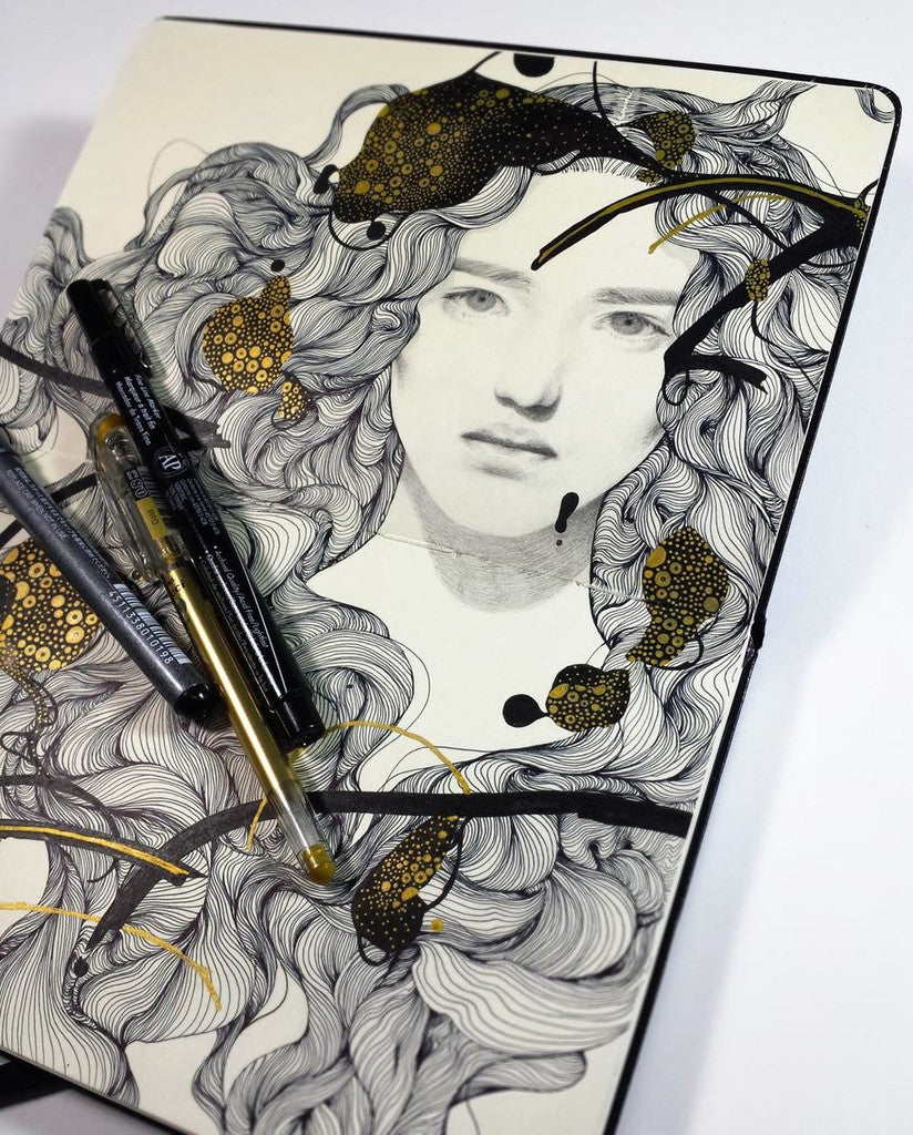 A sketchbook page features a pencil drawing of a womans face, with swirling hair patterns reflecting Tran Nguyens elegance. Black and gold ink adds an abstract touch akin to The Door-Makers Daughter, complemented by two pens on the page—one black and one gold.
