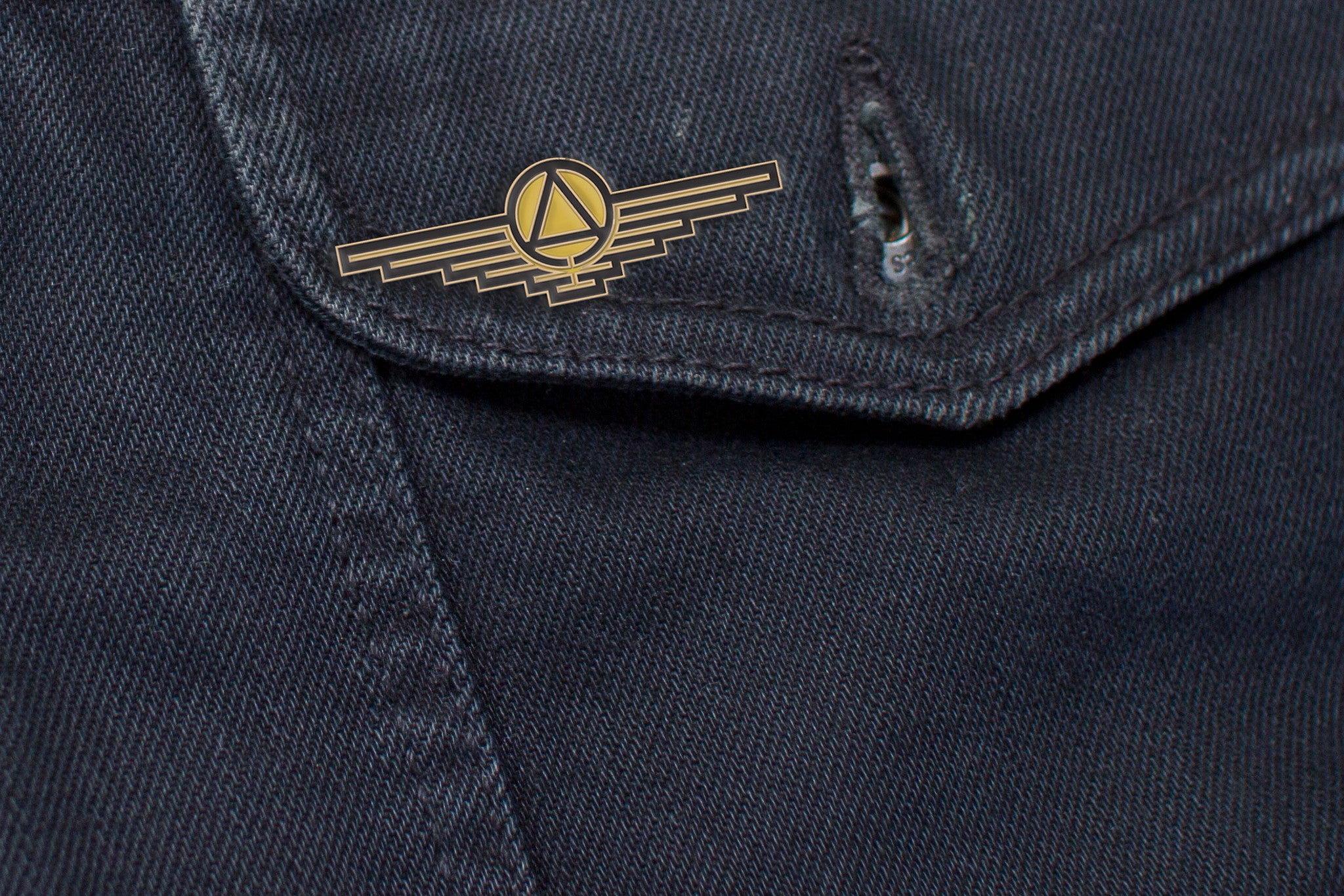 A close-up of dark denim from Joel Daniel Phillips shows a small, gold pin featuring a winged emblem and circular design akin to Red Risings Gold class, securely fastened on a pocket flap.