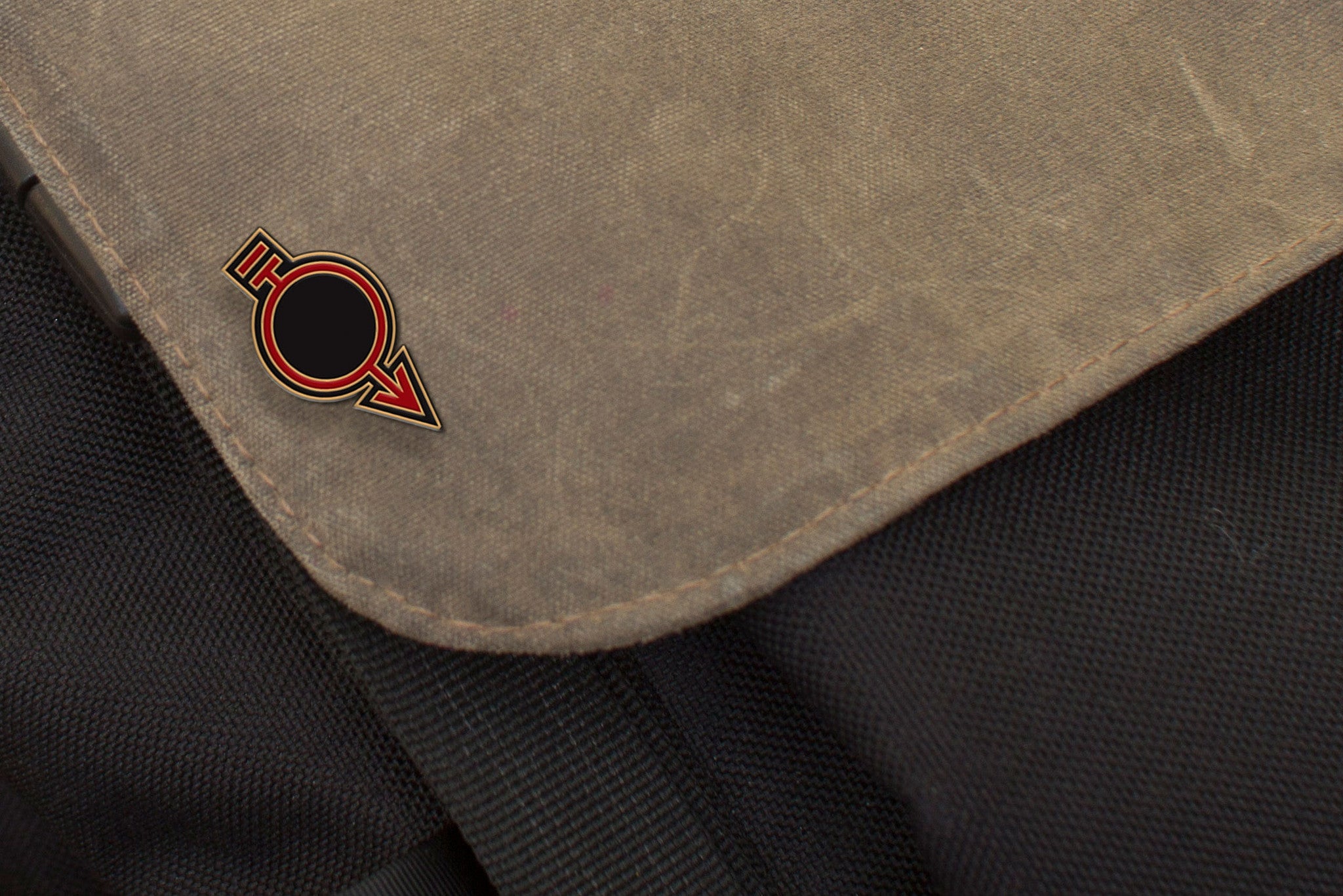 Close-up of a brown fabric flap adorned with an embossed metal pin featuring a black and red circular emblem. The flap rests on black textured material, part of a Joel Daniel Phillips Red Rising-themed bag or backpack.