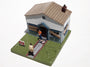 A small Leon Reid IV model features a rundown house with boarded windows, a damaged roof, two figures outside—one holding a baby reminiscent of Welcome Home Soldier—and a red For Sale sign on the green base.
