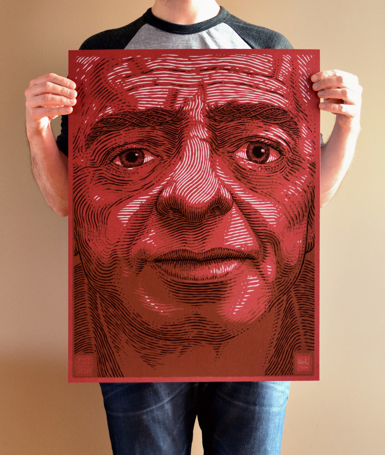 A person in a black and gray shirt holds an eye-catching 3-colour screenprint poster by Bartosz Kosowski. The artwork, possibly of the Man From Another Place, features intricate lines on a large red-toned canvas, capturing a detailed illustration of a mans face.