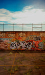 A brick wall with colorful graffiti under a partly cloudy sky resembles a vibrant oil painting. The old concrete ground, dotted with grass and yellow flowers, echoes the style of Jessica Hess. Above the wall, a chain-link fence evokes themes from Alcatraz Redux.