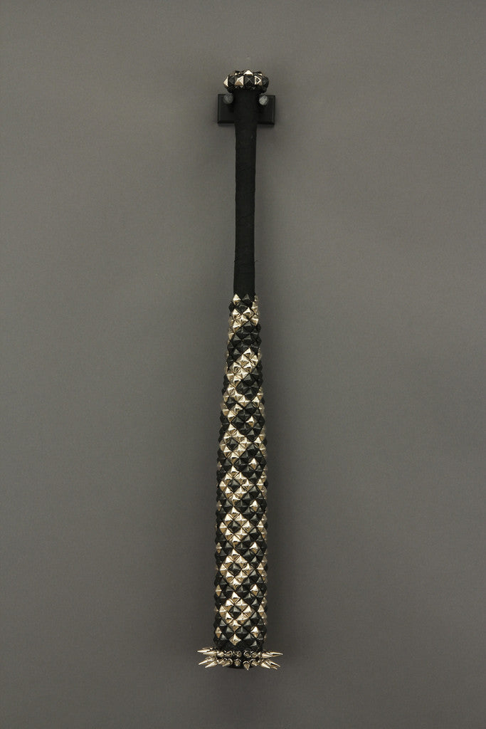 A baseball bat featuring black and white crystals and metallic spikes stands upright against a gray background. The patterns on the handle create a unique visual effect, echoing Lucien Shapiros mixed media artistry.