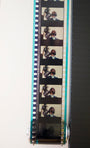 Close-up of a film strip with frames of a person at a mic, mounted vertically, partly unrolled, and displaying sprocket holes. Reminiscent of Kubricks iconic style, it evokes the charm associated with Steve Yamanes designs.