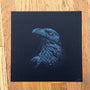 Tracie Chings detailed monochrome drawing of an eagle in profile, highlighting its feathers and sharp beak, is set against a black background. The artwork rests on a wooden surface, ideal for custom framing.
