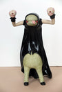 A green creature with a large belly and a black cloak, sculpted by Doubleparlour from resin, sticks its tongue out. It has two raised arms ending in mask-like faces and stands on two legs with black shoes, detailed with acrylic paint.