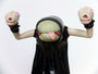 A cartoonish creature from Doubleparlour features a green face, red eyes, a protruding tongue, and a black hooded robe. Its arms extend upward, each with a head having closed eyes and an open mouth—hand-sculpted resin enhances its quirky design.