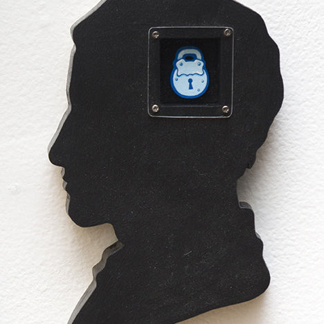 A hand-cut wood plaque by Matt Ritchie features a persons silhouette with an acrylic painting inside, depicting a cartoonish blue padlock icon in a square frame.
