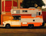 A retro Citrus Camper, with horizontal stripes and a raised cabin reminiscent of Jessica Hesss oil on canvas art, is parked against red and beige walls and lush green leaves.
